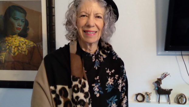 An older white woman in a colorful jacket and beret poses, smiling, for a picture in an eclectically decorated living room. She evinces style and joie de vivre.