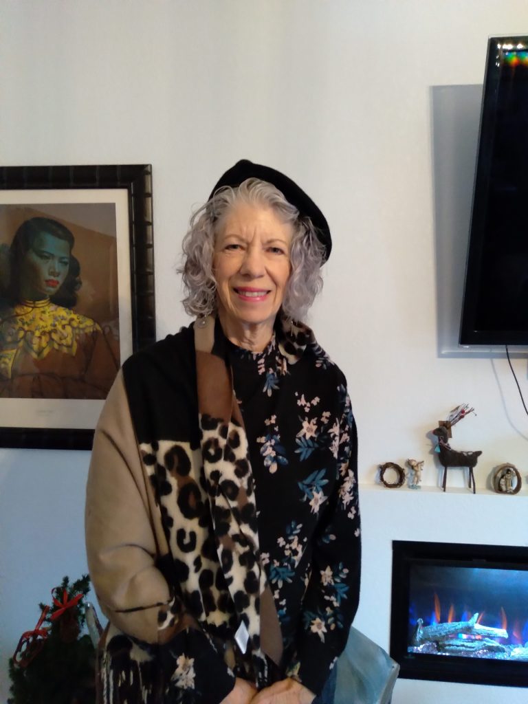 An older white woman in a colorful jacket and beret poses, smiling, for a picture in an eclectically decorated living room. She evinces style and joie de vivre.
