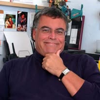 A sturdy looking middle aged white man smiles mischieviously at the camera with his hand on his chin, sitting in what is presumably his study. He looks confident and at ease.