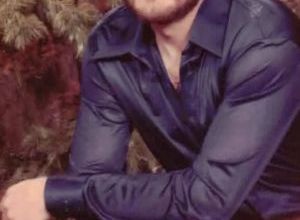 A Caucasian man in a blue shirt and brown pants rests his elbows on his knees in a relaxed studio portrait. He has brown hair and a brown beard.
