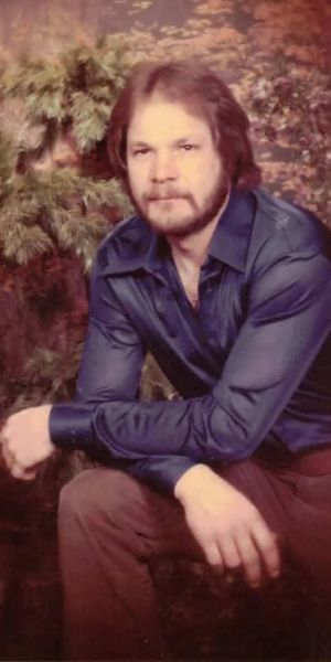 A Caucasian man in a blue shirt and brown pants rests his elbows on his knees in a relaxed studio portrait. He has brown hair and a brown beard.