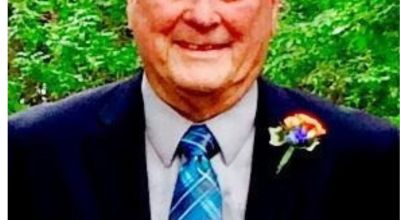 Portrait of Frank L Servoss, an older white gentleman in a dark blue suit with a light blue plaid tie. He is wearing a pink boutineer and smiling, it appears to be a celebratory occasion.