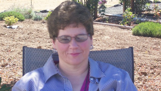 A smiling white woman with short brown hair, glasses sits in the shade outside on a lawn chair. She smiles knowingly.
