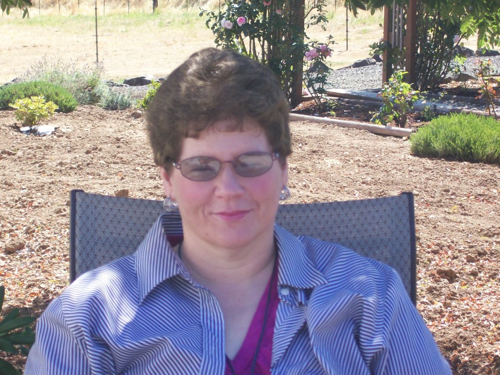 A smiling white woman with short brown hair, glasses sits in the shade outside on a lawn chair. She smiles knowingly.