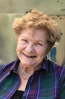 An older white lady with red hair wearing a green and purple checked shirt beams at the camera joyfully