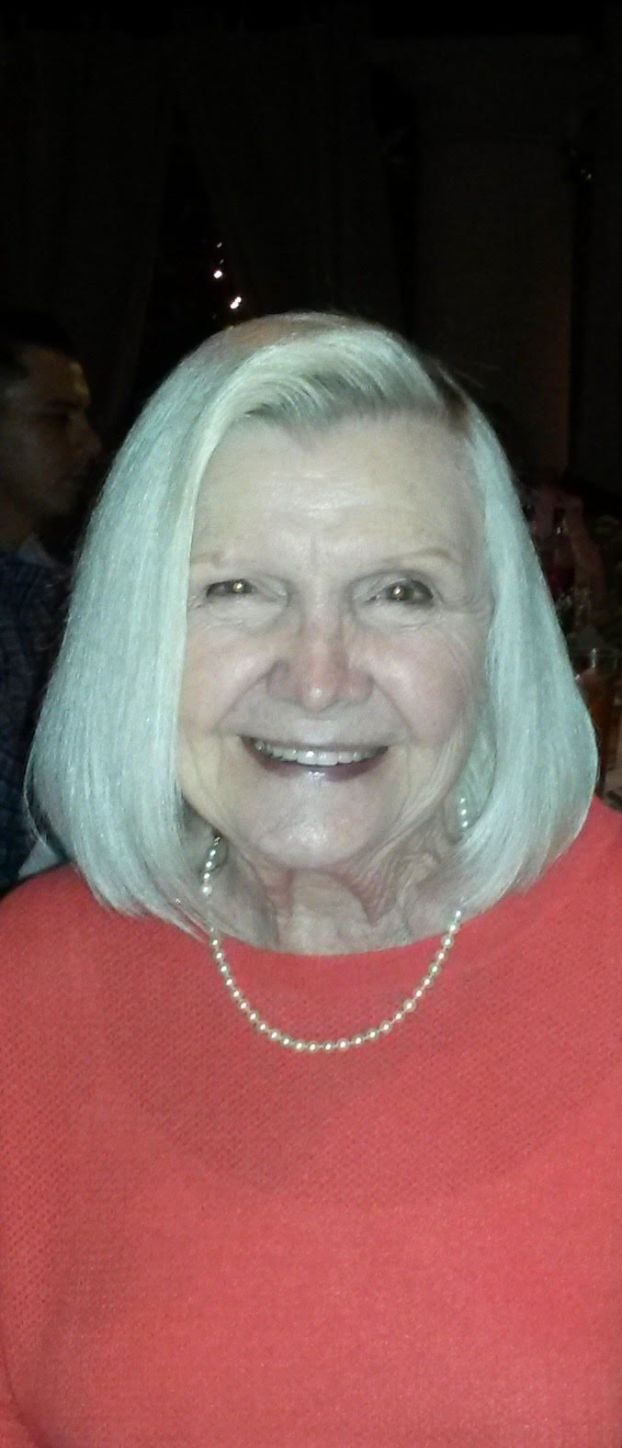 An old white woman with white hair wearing pearls and a red dress smiles for the camera. She looks very contented.