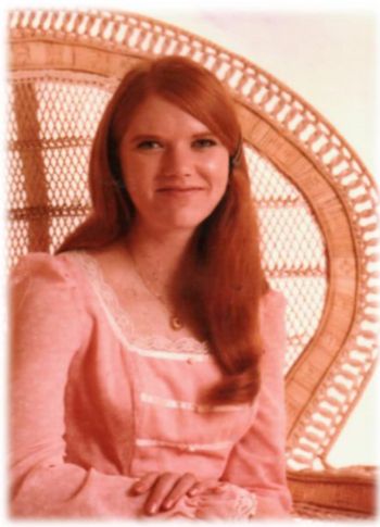 A red haired woman smiles at the camera. She's wearing a pink gown and is resting her hands in her lap as she sits in a big wicker chair. She's quite lovely.