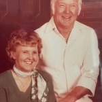 Mae and Rod Boren, a married couple, a woman sits for a photo in a green sweater, she had red hair and smiling with happy eyes. Rod is holding her hand in a white shirt, looking happy.