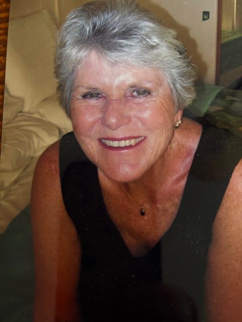 An older white lady with short gray hair and a black tanktop smiles at the camera. She looks vivacious and happy.