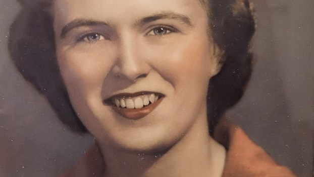 An old colorized photo of a young white woman with auburn hair wearing a brown sweater and smiling for her portrait.