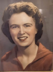 An old colorized photo of a young white woman with auburn hair wearing a brown sweater and smiling for her portrait.