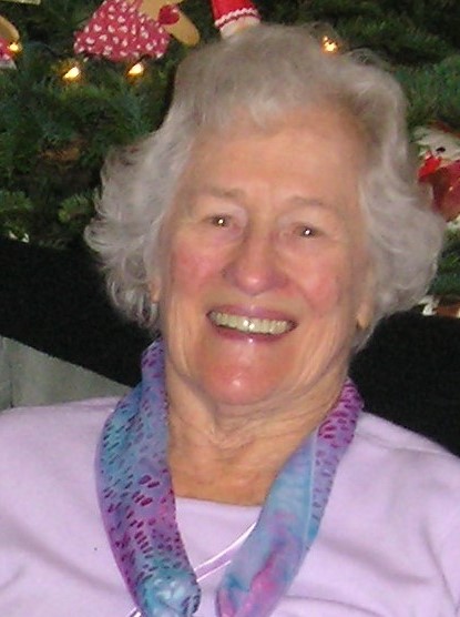 An older white woman smiles for the camera in a purple sweater with a blue and magenta scarf. She looks very happy.