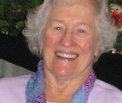 An older white woman smiles for the camera in a purple sweater with a blue and magenta scarf. She looks very happy.