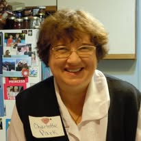 A smiling older lady with glasses and short Auburn hair, wearing a name tag, she looks like a grade school teacher.