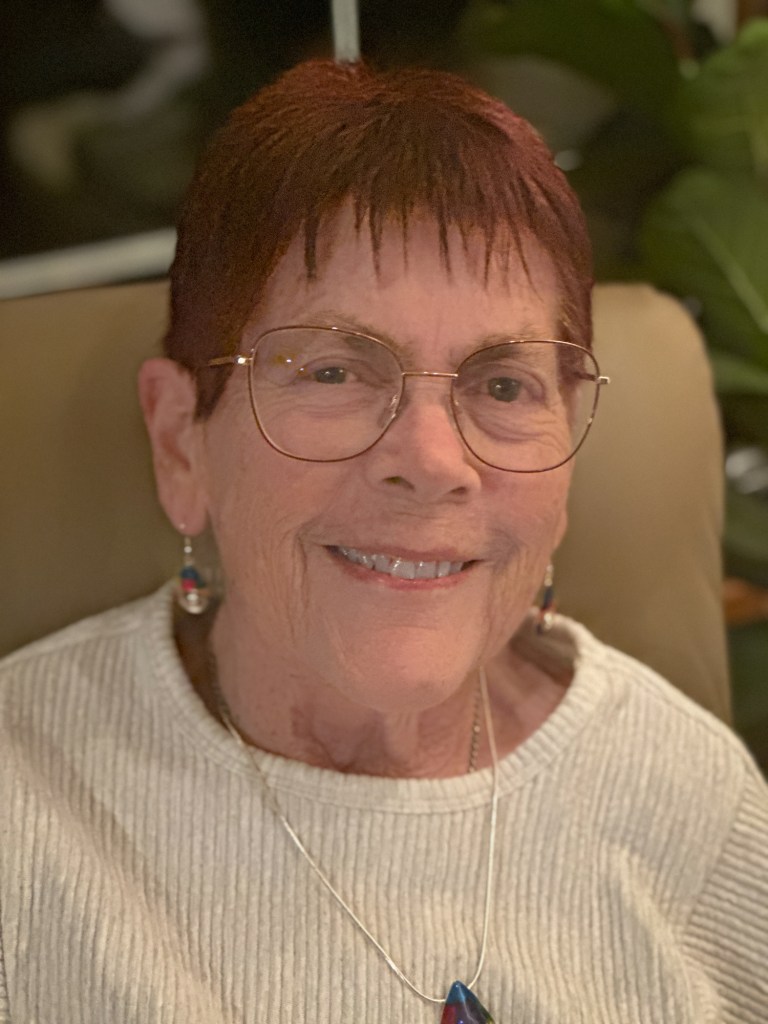 An older white lady with bright red hair and big round glasses is sitting and smiling for the camera wearing a white sweater. She looks happy.