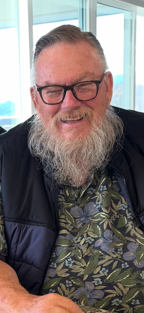 A smiling older white man with black framed glasses and a white beard looking like a biker santa claus beams for the camera, he does indeed look very jolly.