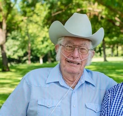 An elderly white man wearing a cowboy hat and blue shirt, glasses and fluffy white muttonchops smiles for the camera. He looks like a jolly sort of fella.