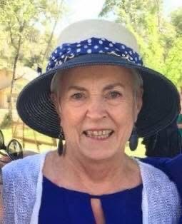 An older white lady with a purple blouse, white shall, and white and purple hat smiles for the camera. She looks friendly.