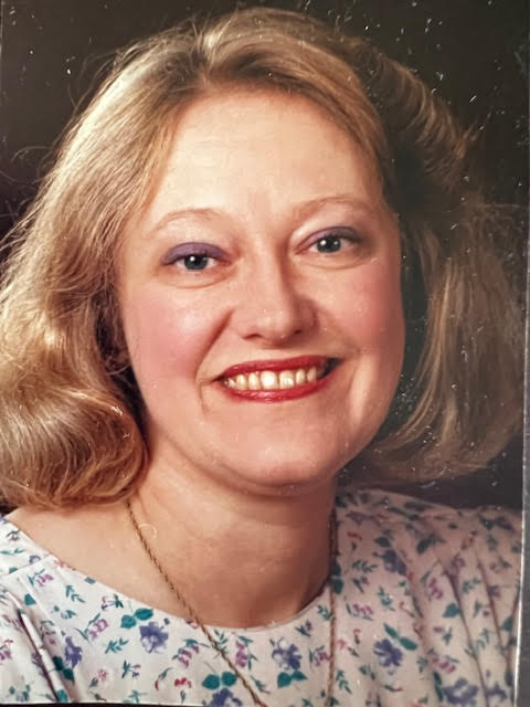 A blonde woman with bright red lipstick and a floral blouse smiles for the camera.