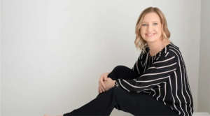 A blond woman in a black and white striped blouse with black trousers poses casually for the camera.
